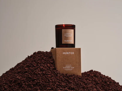 Hunter coconut sunscreen sand scented candle on box, displayed on brown textured rocks